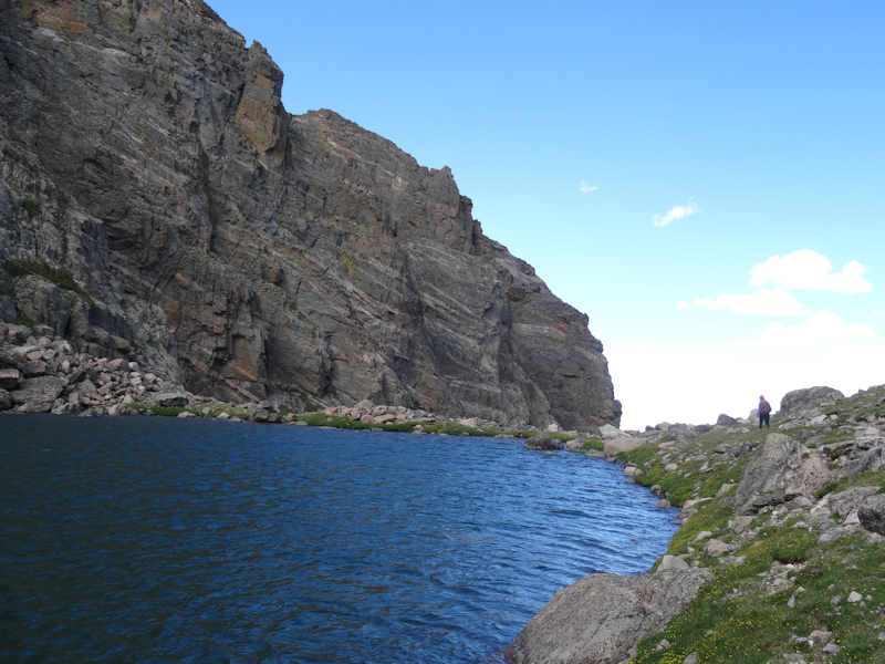 a far-away person stands to the right side of a body of water under a cliffside